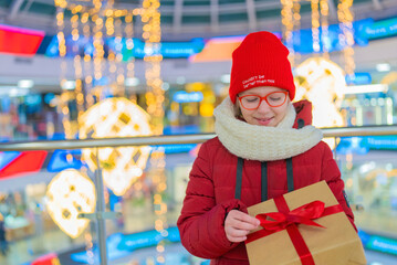 teenage girl in a white jacket in a mall holding a box with a gift on the background of a shop window with discounts during a sale