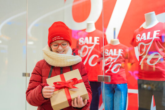 teenage girl in a white jacket in a mall holding a box with a gift on the background of a shop window with discounts during a sale