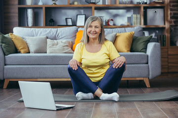 Obraz premium Portrait of active senior woman sitting at home on lotus floor and looking at camera, happy grandmother doing yoga online using laptop