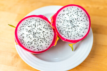 Closeup macro of cut open pink red dragon dragonfruit fruit slices halves half inside flesh cross section with black seeds on white plate and wooden table