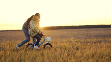 Naklejka na ściany i meble childhood dream. woman mother teaches child to ride bike in the park. Driving lesson for kid from mom at sunset. parent helps child to keep balance and pedal. Kid learns steer bike sun. happy family