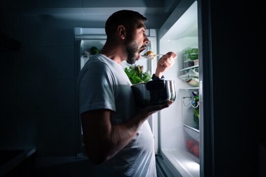 Hungry man eating food at night from open fridge. Man taking midnight snack from refrigerator