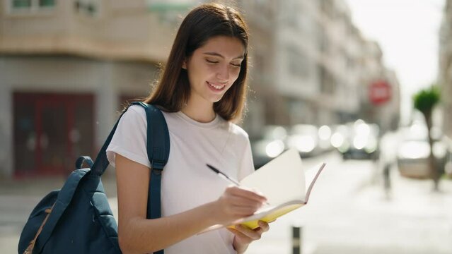 Young hispanic girl student smiling confident writing on book at street