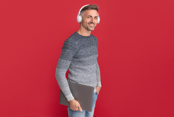 Happy man in casual listen to audio course in headphones holding laptop red background, e-learning