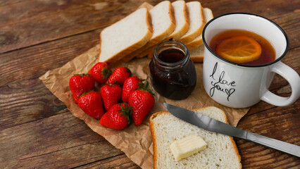 White cup of black tea with lemon. Sandwiches or toasts with butter. Strawberry and jam jar on wooden table. Breakfast and happy morning concept