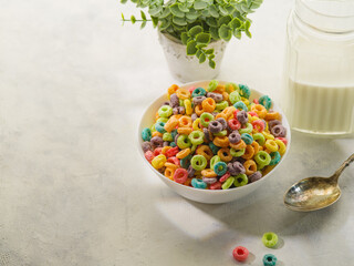 A bowl with multi-colored grain fruit rings, a spoon, milk in a jug and a decor - an indoor flower on a white background. Quick whole grain breakfasts, baby food, vitamins, antioxidants.