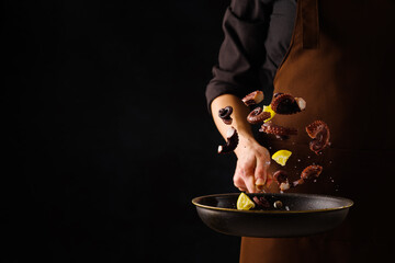 A professional chef in a dark uniform cooks octopus with lemon slices in a frying pan on a black...