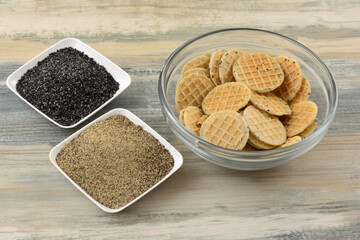 Black pepper and poppy seed wafer cookies in glass bowl with poppy seeds and black pepper on two small ingredient dishes