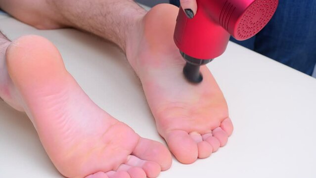 Hand of unrecognized female masseur with electric percussion vibratory massager massages the soles of the athlete's feet. Sports body regenerating massage therapy, close-up view