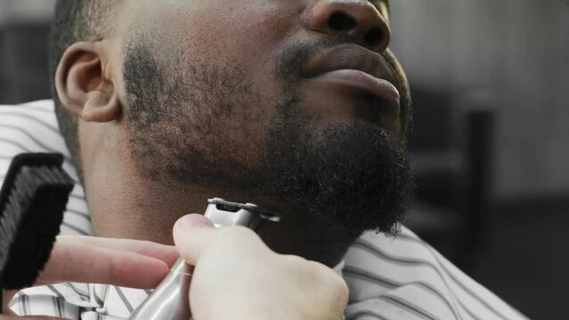 Close-up. An African American man sits in a barber chair and has his beard shaved off. Salon. barbershop services
