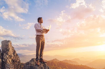 Businessman working outdoors with laptop computer standing on top rock mountain at sunset...