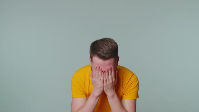 Upset disappointed handsome stylish man in yellow t-shirt wipes tears and cries from despair, being sad because of unfair things. Young adult guy. Indoors studio shot isolated alone on gray background