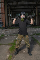 A soldier or partisan with an ak-74 in a modern gas mask, black helmet, T-shirt and camouflage pants stands among the ruins of destroyed buildings