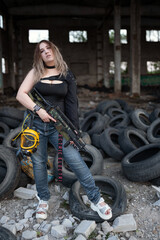 Fototapeta na wymiar A beautiful strong girl with big breasts stands with a machine gun and a gas mask on her hip among the ruins of a destroyed building and a pile of old tires. Guerrilla, rebel or resistance fighter