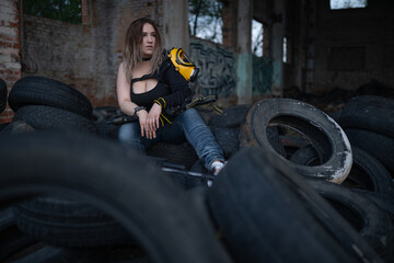 Fototapeta na wymiar A beautiful strong girl with big breasts sits with a machine gun and a gas mask on her hip the ruins of a destroyed building and a pile of old tires. Guerrilla, rebel or resistance fighter