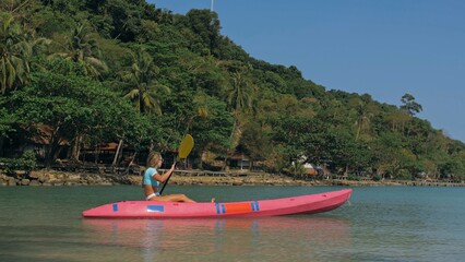 Young blonde woman in blue swimsuit rows pink plastic canoe along azure sea bay past island with palms under blue sky at resort. Traveling to tropical countries. Girl is sailing on kayak in ocean.