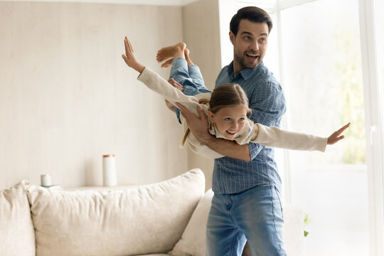Happy dad lifting and rocking excited daughter girl playing airplane with flying open hands, holding little kid in arms, having fun, enjoying active exercises. Family home entertainment concept