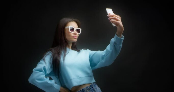 Brunette with long hair in white sunglasses and a blue sweater takes a selfie on her phone. Young Asian woman makes faces in front of a smartphone taking stupid selfies isolated on a black background.