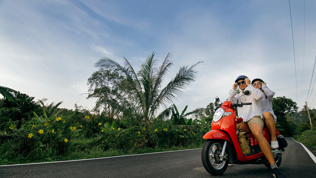 Love couple on red motorbike in white clothes to go on forest road trail trip. Two caucasian tourist woman man drive on scooter. Motorcycle rent, safety helmet, sunglasses. Asia Thailand ride tourism.