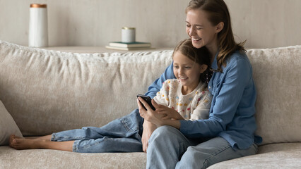 Happy laughing mom and smiling kid using online ecommerce app on smartphone, shopping on internet together, watching funny video, media content on mobile phone, relaxing on sofa at home