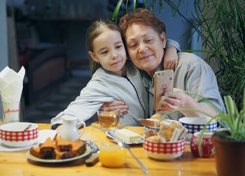 Grandma and granddaughter are sitting in the kitchen, looking at photos on their phones and taking selfies. Warm family relationships.