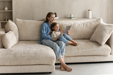 Happy young mom embracing sweet gen Z daughter child on comfortable couch, watching kid using learning app on tablet computer, playing online game, reading book, browsing internet, shopping