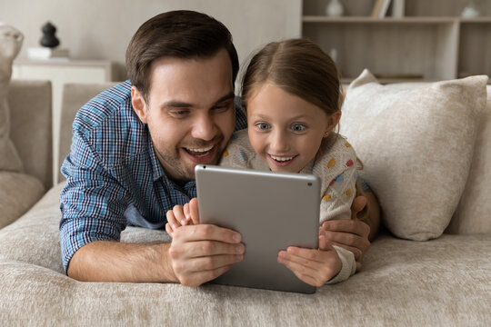 Excited happy millennial daddy and surprised gen Z daughter kid reading book, message, email on tablet computer, watching video, online movie, media on internet, getting good amazing great news