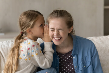 Cute kid telling secret to happy laughing elder sister, nanny, daycare teacher, whispering in ear. Pretty loving daughter girl and young mom enjoying friendship, having fun, talking at home - obrazy, fototapety, plakaty