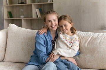 Happy young pretty mother embracing sweet daughter kid with love, gratitude head shot portrait. Mom and child sitting on couch in living room, hugging, smiling, looking at camera. Motherhood concept