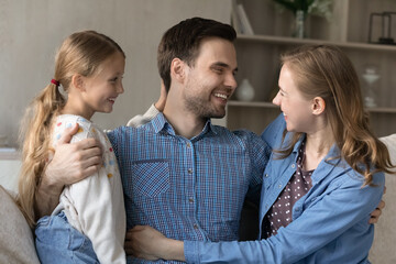 Happy young wife and cute little daughter embracing cheerful grateful husband and father at home. Family couple of parents and kid hugging on sofa at home with love, care, enjoying closeness, unity