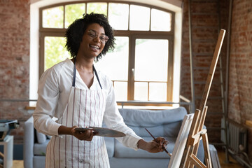 Portrait of happy creative young African American curly woman in eyeglasses wearing apron, mixing...