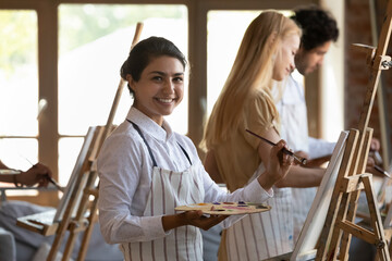 Portrait of beautiful millennial inspired Indian woman in apron holding palette with mixed oil paints, enjoying drawing on easel at group lesson in modern loft studio, enjoying creative hobby activity