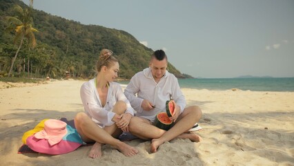 Fototapeta na wymiar The cheerful love couple holding and eating slices of watermelon on tropical sand beach sea. Romantic lovers two people caucasian spend summer weekend in outdoor. Hat, backpack white shirt beachwear.