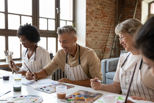 Happy Handsome Man In Apron Drawing Picture With Paints On Paper, Enjoying Creative Art Master Class Workshop With Smiling Multiracial Young And Old People, Sitting Together At Table In Loft Studio.