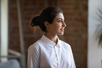 Fototapeta premium Head shot motivated happy young female Indian business owner employee worker looking in distance, thinking of career opportunities or professional challenges, standing alone in modern workplace.