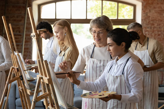 Skilled happy old female teacher helping young Indian woman drawing pictures at easel, giving advices about creative artwork, educating at group lesson with diverse people in modern loft studio.