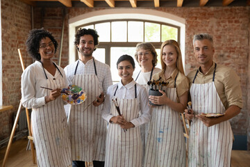 Group portrait of smiling multiracial people of different generations in painted aprons holding set...