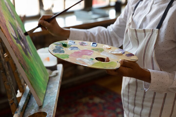 Close up cropped young male artist holding palette with mixed acrylic paints, involved in creating artwork on canvas, improving art painting skills, enjoying hobby activity in modern studio or home.