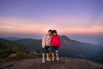 Obraz premium Two cute little girls hugging on top of a mountain and looking at the camera against a sunset background. Hiking adventures with family on vacation.