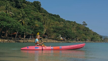 Young blonde woman in blue swimsuit rows pink plastic canoe along azure sea bay past island with palms under blue sky at resort. Traveling to tropical countries. Girl is sailing on kayak in ocean.