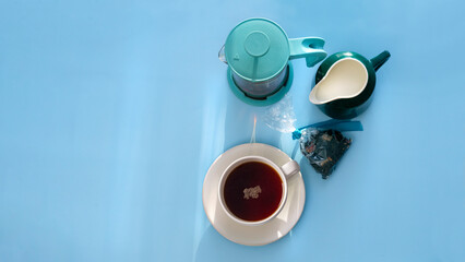 A cup of black hot tea with milk in jug and teapot stand on blue table with sunbeams. View from above.