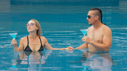 The loving couple hugs and kisses, drinking blue cocktail alcohol liquor in swimming pool at hotel...