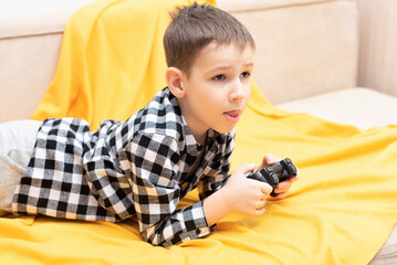 The child boy in checked shirt lying on the couch with black joystick in his hands playing the...