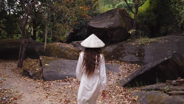 Young european woman traveler in long white dress and vietnamese hat walking in forest near huge boulders, big stones in Vat Phou – ruined Khmer Hindu Temple. Dry leaves, green trees around. Slow