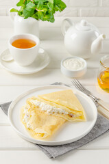 Traditional thin pancakes or crepes with cottage cheese and raisins, honey and sour cream on a white wooden background. Food for Maslenitsa. Copy space.