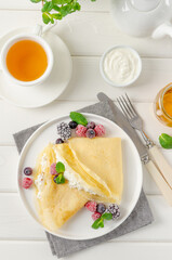 Traditional thin pancakes or crepes with cottage cheese and raisins with berries, honey and sour cream on a white wooden background. Food for Maslenitsa. Copy space.