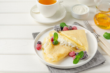 Traditional thin pancakes or crepes with cottage cheese and raisins with berries, honey and sour cream on a white wooden background. Food for Maslenitsa. Copy space.