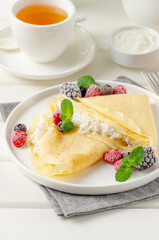 Traditional thin pancakes or crepes with cottage cheese and raisins with berries, honey and sour cream on a white wooden background. Food for Maslenitsa. Copy space.
