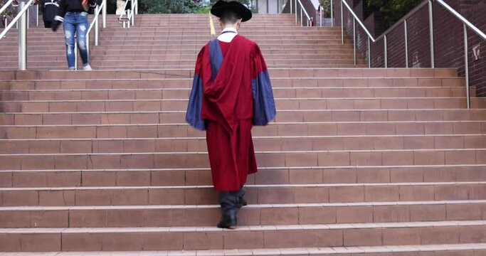 young male PhD graduate student in red academic dress walk up in stairway in university campus during congregation day, concept of success or Moving Up the Career Ladder