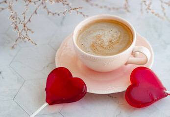 Coffee in a pink ceramic cup surrounded by white flowers, with a red heart-shaped lollipop on top. Delicate breakfast in a stylish interior in a light key
